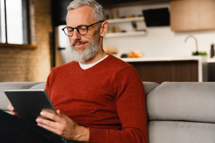 Closeup portrait of a smiling caucasian middle-aged mature man in glasses using digital tablet for e-learning, paying bills online, e-commerce, remote work, social media online at home