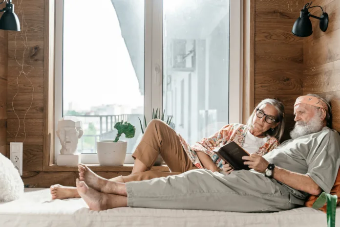 Elderly man lying on bed reading a book beside an elderly woman