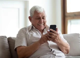 eSamonaročanje: naročite se na obisk na upravni enoti Happy older man sitting on comfortable sofa at home, using smartphone. Pleasant 80s grandfather reading sms with good news, typing message for grown up kids, shopping in mobile app, watching photos.