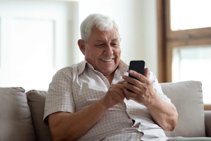 Happy older man sitting on comfortable sofa at home, using smartphone. Pleasant 80s grandfather reading sms with good news, typing message for grown up kids, shopping in mobile app, watching photos.