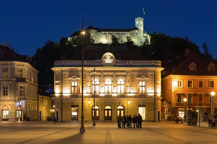 Ljubljana, Slovenia - October 4, 2018: The Slovenian Philharmonic Building on the background of Ljubljana Castle in dusk. The building was constructed in 1891 by design of the Austrian architect Adolf Wagner. The castle was founded in the 11th century.