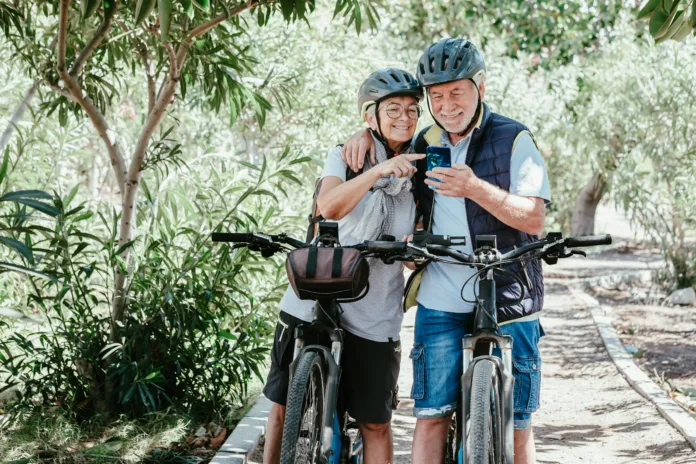 Active senior caucasian couple with electro bikes looking at smart phone outdoors in the park. Two happy elderly people enjoying nature and healthy lifestyle