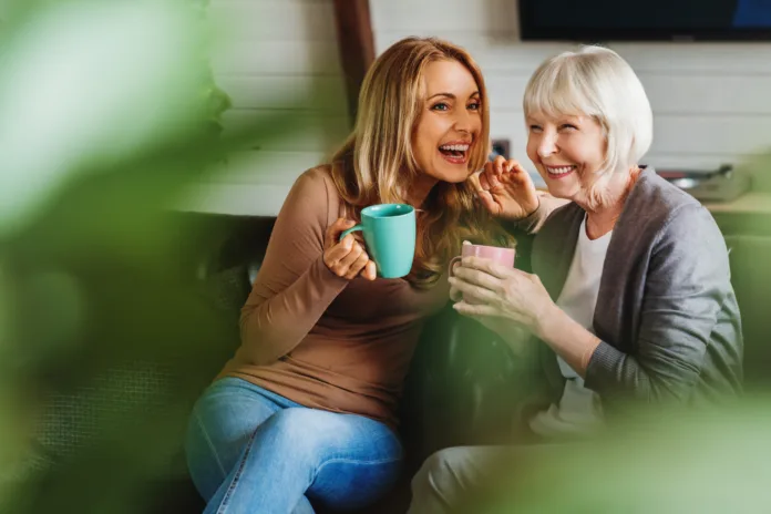 Happy senior mother with adult daughter sitting on couch and holding cups with coffee or tea at home. Togetherness concept