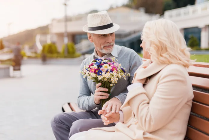 Two pensioners are sitting on a bench in the alley. An elderly man gives a woman flowers. She is delighted with the gift