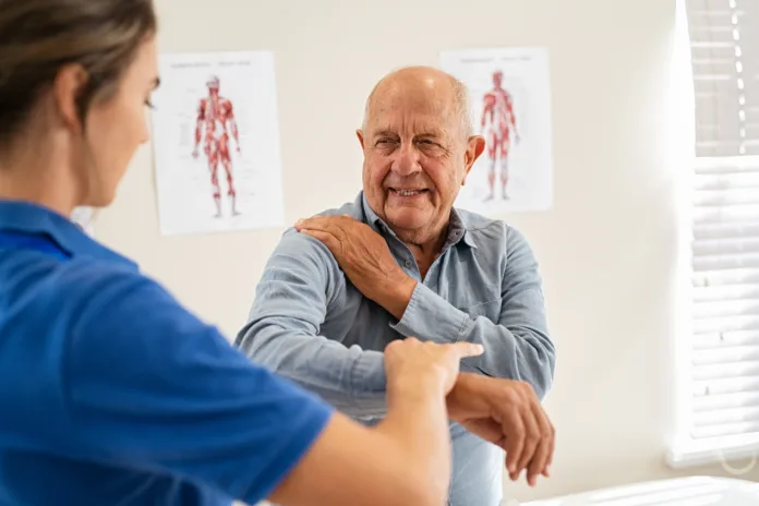 Young woman physiotherapist helping senior man with elbow exercise in clinic. Young woman doctor checking elbow of senior patient. Old man during an appointment with professional osteopath in private clinic working and massaging his shoulder pain.