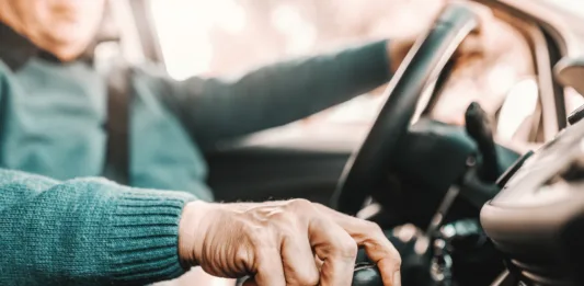 Close up of senior man holding one hand on gearshift and other on steering wheel while sitting in his car. Selective focus on hand.