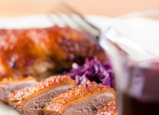 3 preprosti recepti za popolno Martinovo večerjo Close-up selective focus of roast duck sliced into pieces on a white plate behind a red glass of wine. The plate rests on a light-brown wooden table. On the plate behind the duck is shredded red cabbage, with an unfocused red and gold food item behind that. Fork tines are visible near the back of the plate.