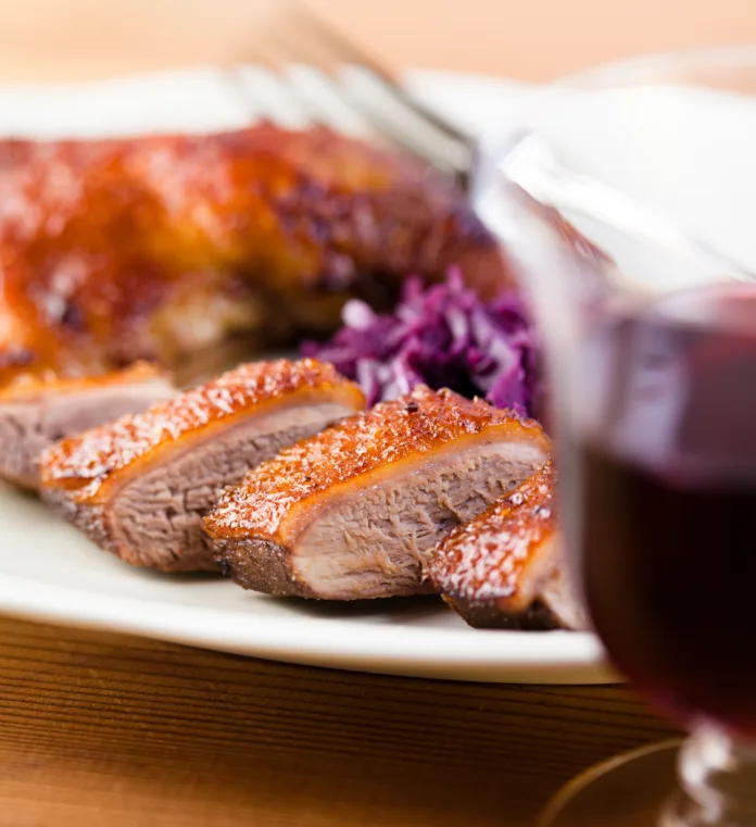 Close-up selective focus of roast duck sliced into pieces on a white plate behind a red glass of wine.  The plate rests on a light-brown wooden table.  On the plate behind the duck is shredded red cabbage, with an unfocused red and gold food item behind that.  Fork tines are visible near the back of the plate.