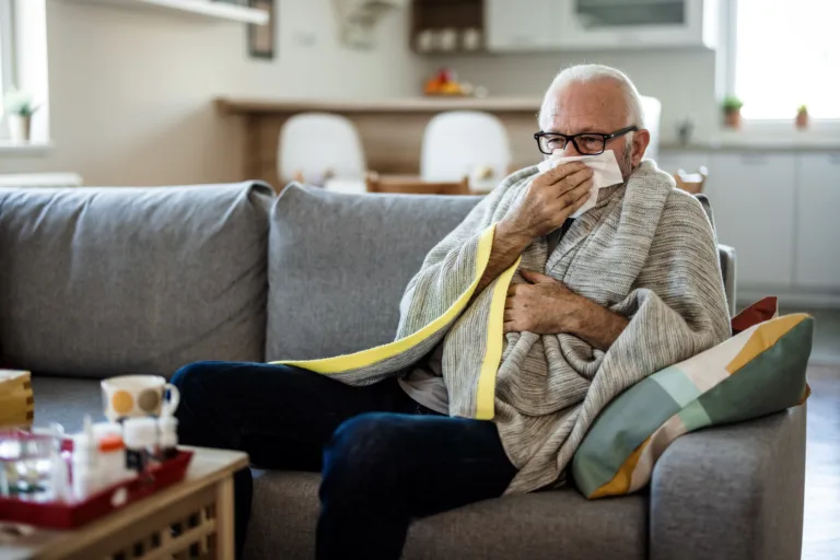 Senior man at home suffering with cold or flu virus. Shot of a senior man blowing his nose with a tissue at home.