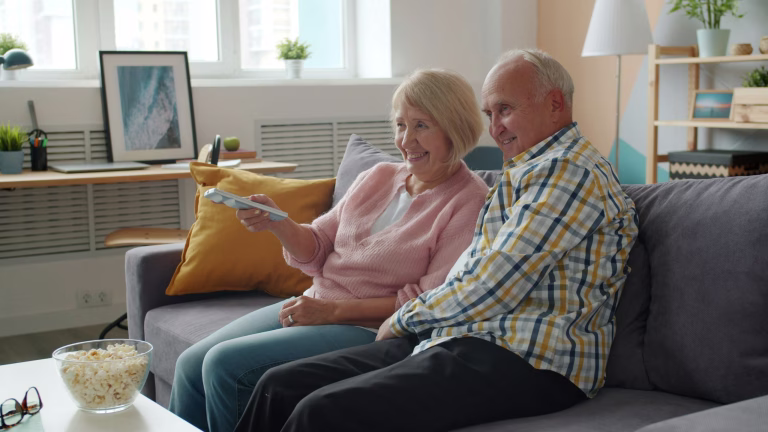 elderly man and woman watching TV laughing talking enjoying movie at home on sofa. Entertainment, lifestyle and leisure concept.