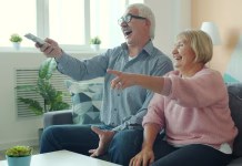 So resničnostni šovi sploh dobra izbira za ogled? Joyful old lady is watching TV with handsome elderly husband laughing having fun enjoying movie at home. Entertainment, people and lifestyle concept.