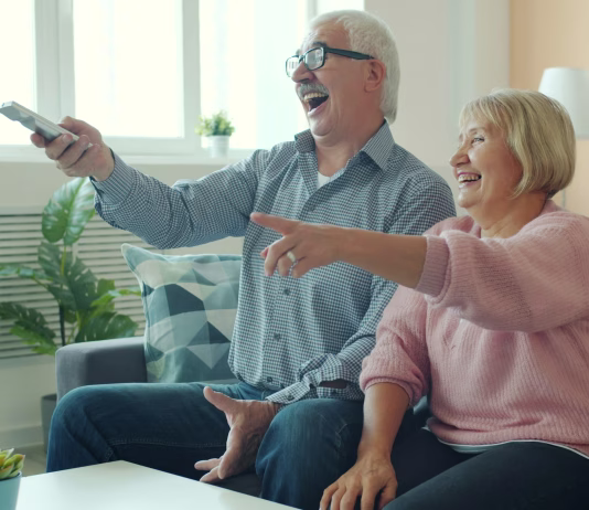 So resničnostni šovi sploh dobra izbira za ogled? Joyful old lady is watching TV with handsome elderly husband laughing having fun enjoying movie at home. Entertainment, people and lifestyle concept.