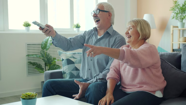 Joyful old lady is watching TV with handsome elderly husband laughing having fun enjoying movie at home. Entertainment, people and lifestyle concept.