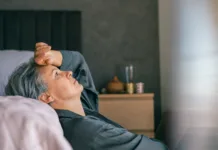 Stalna utrujenost: kdaj k zdravniku? A mature woman with gray hair lies in bed, looking exhausted and thoughtful, reflecting midlife stress or menopause challenges in a calm, intimate home environment.