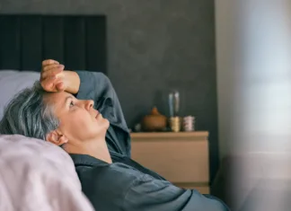 Stalna utrujenost: kdaj k zdravniku? A mature woman with gray hair lies in bed, looking exhausted and thoughtful, reflecting midlife stress or menopause challenges in a calm, intimate home environment.