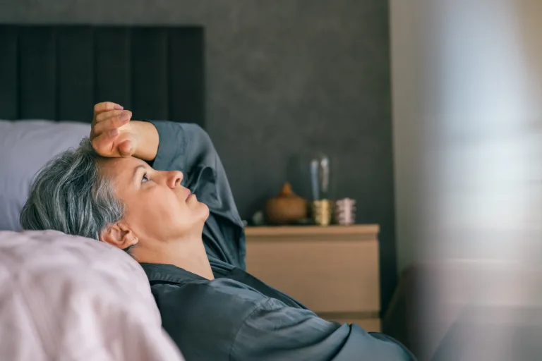A mature woman with gray hair lies in bed, looking exhausted and thoughtful, reflecting midlife stress or menopause challenges in a calm, intimate home environment.