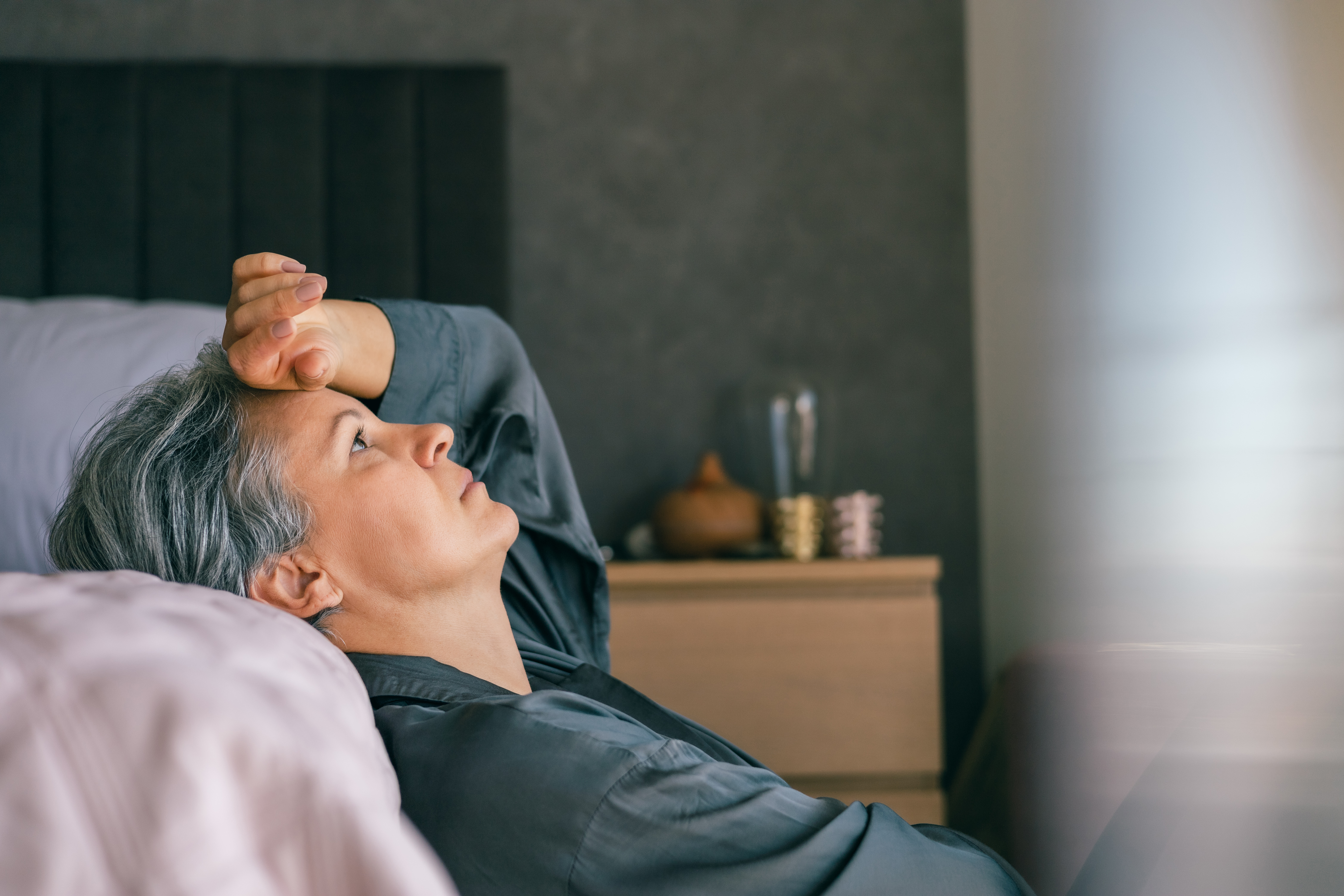 Stalna utrujenost: kdaj k zdravniku? A mature woman with gray hair lies in bed, looking exhausted and thoughtful, reflecting midlife stress or menopause challenges in a calm, intimate home environment.