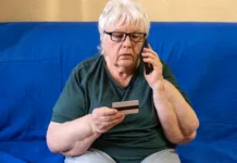 Prevaranti računajo na to napako: tako zaščitite telefon in prihranke Serious old woman in glasses sitting on sofa holding credit card and talking on phone. Concept of scam, money loss, banking protection, finance fraud, customer service, remote payment.