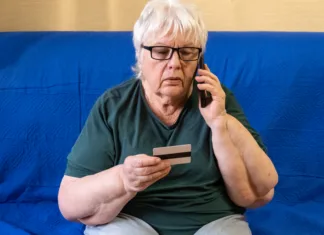 Prevaranti računajo na to napako: tako zaščitite telefon in prihranke Serious old woman in glasses sitting on sofa holding credit card and talking on phone. Concept of scam, money loss, banking protection, finance fraud, customer service, remote payment.