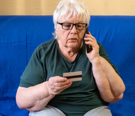 Prevaranti računajo na to napako: tako zaščitite telefon in prihranke Serious old woman in glasses sitting on sofa holding credit card and talking on phone. Concept of scam, money loss, banking protection, finance fraud, customer service, remote payment.
