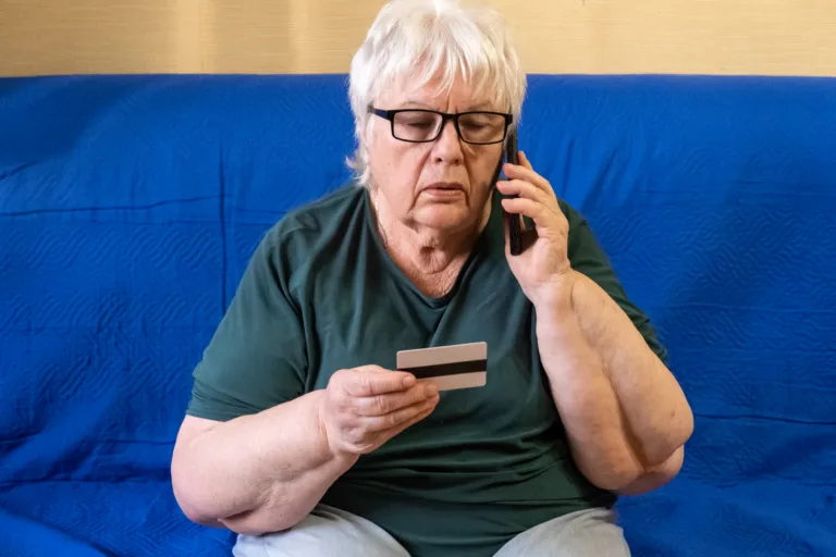 Serious old woman in glasses sitting on sofa holding credit card and talking on phone. Concept of scam, money loss, banking protection, finance fraud, customer service, remote payment.