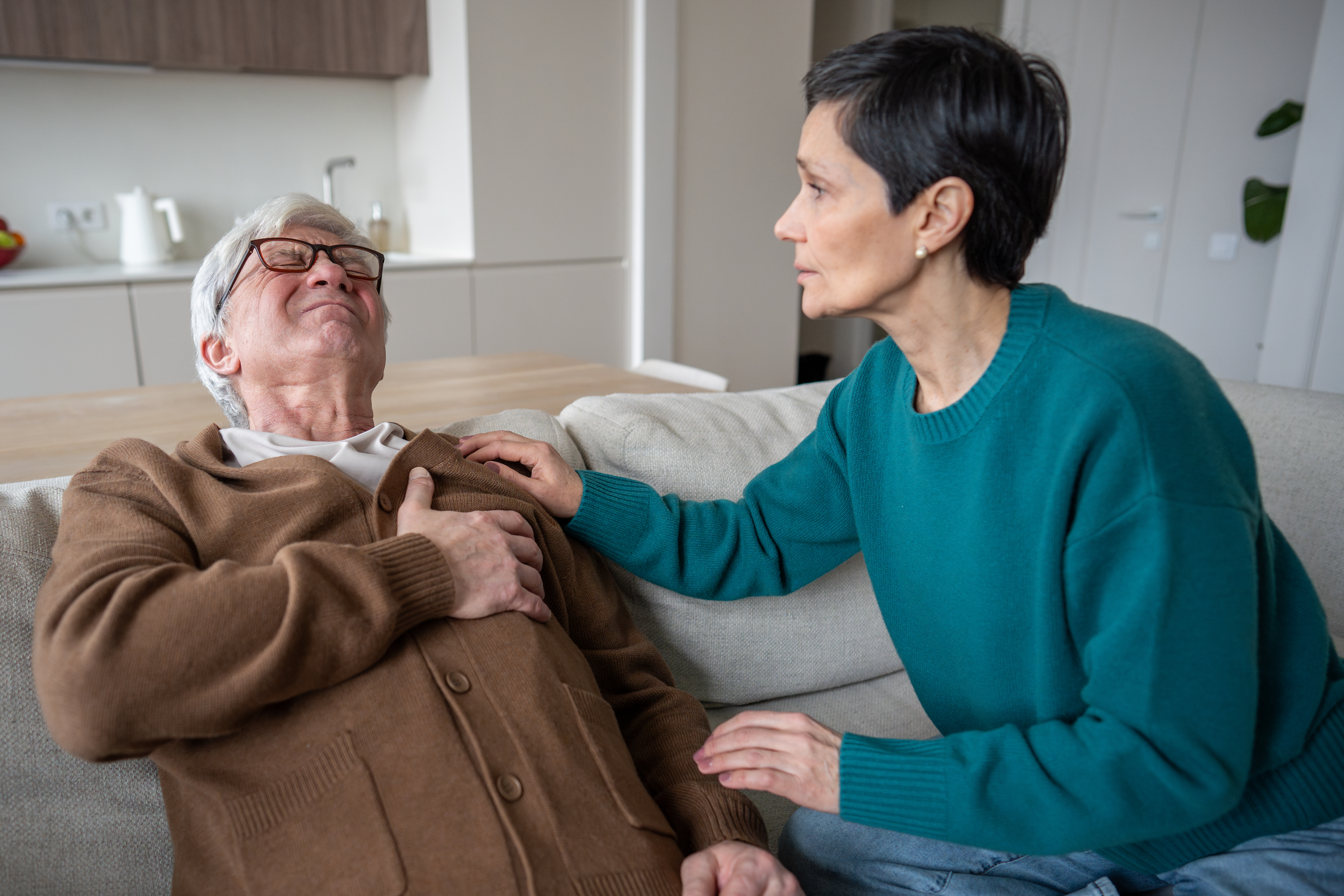 Worried panic wife clings to suffering husband hand during cardiac attack. Physical suffer scared man holding chest heavily breathing, emotional support, mature couple in distress, senior male in pain