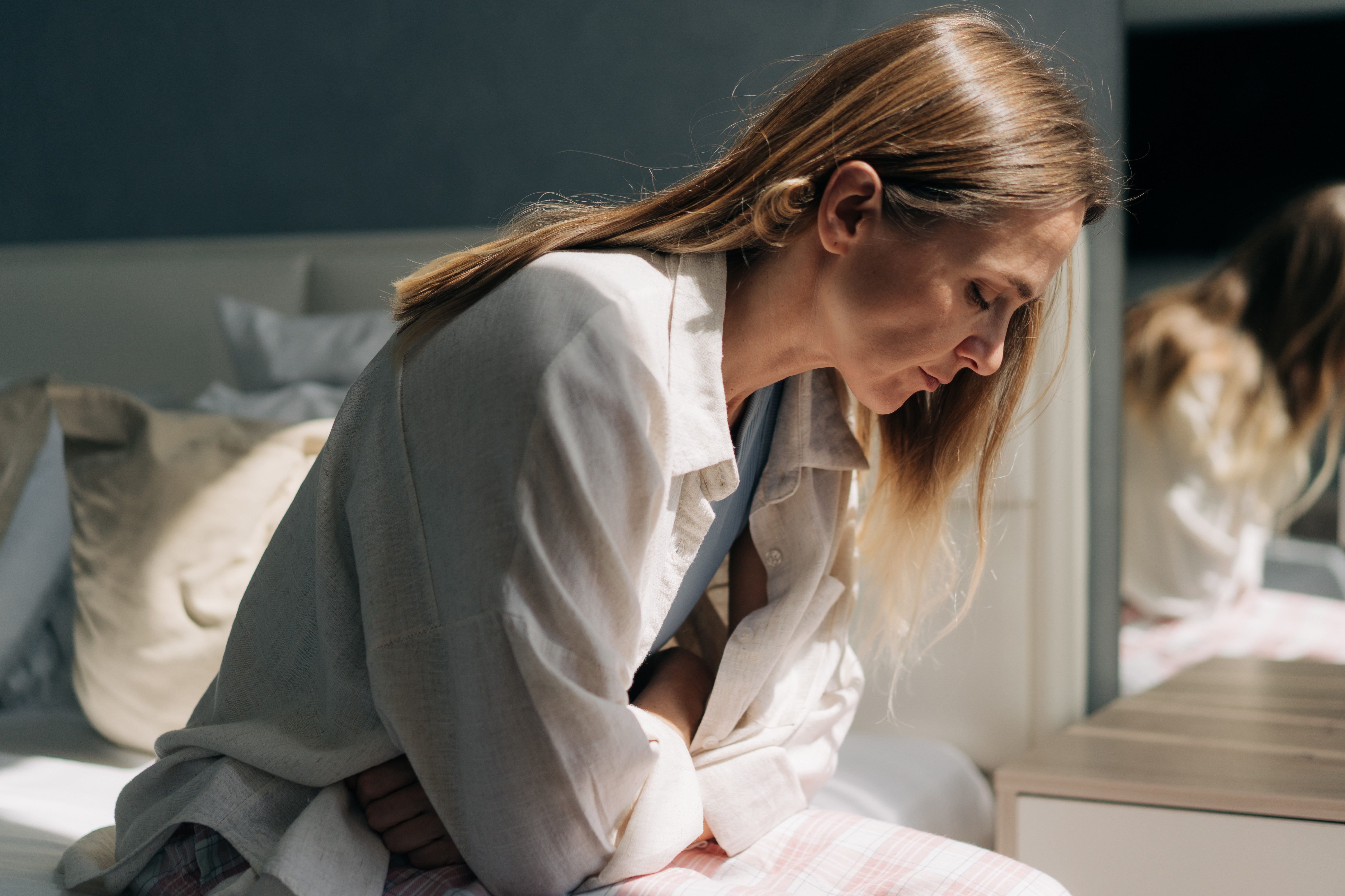 A middle-aged woman sits on the bed and holds her hands on stomach in pain. Menstrual pain, women's health.