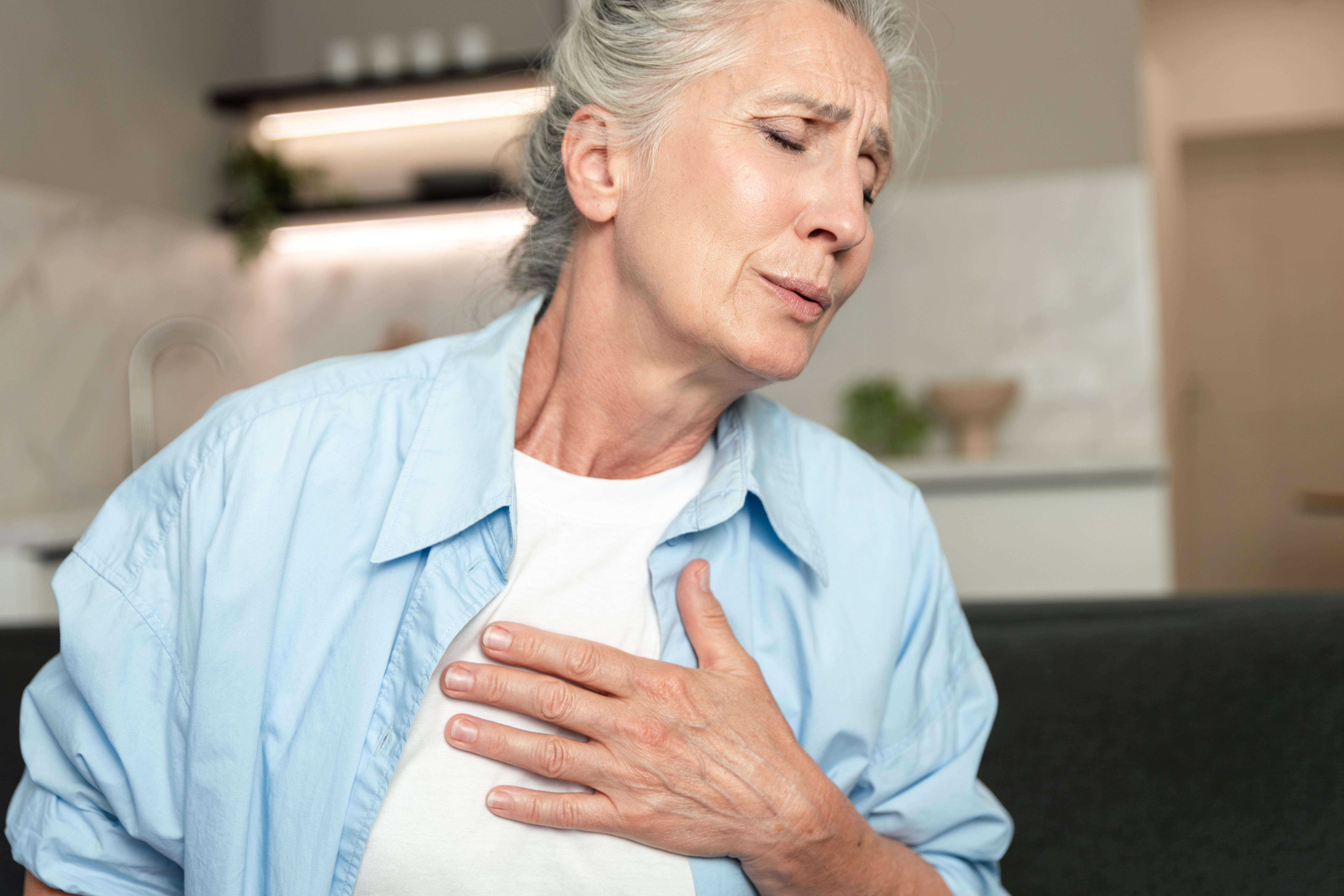Acute heart pain, cardiac disease and chronic illness concepts. Portrait of elderly woman with closed eyes experiencing discomfort, clutching her chest with an exhausted expression.