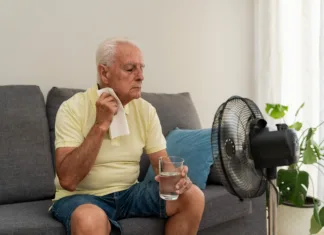Že 1300 mrtvih zaradi vročine. Kako si pomagate, če nimate klimatske naprave? Elderly man refreshing himself with a glass of water and an electric fan, trying to cope with high temperature at home during summer heatwave