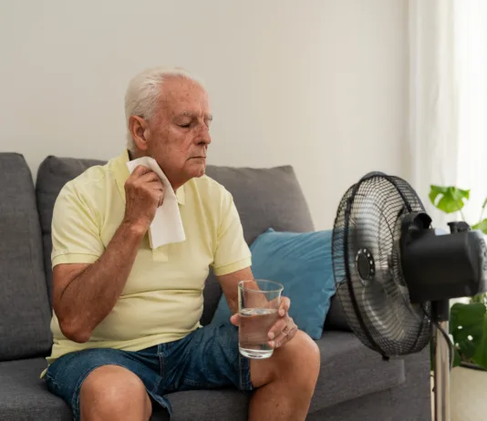 Že 1300 mrtvih zaradi vročine. Kako si pomagate, če nimate klimatske naprave? Elderly man refreshing himself with a glass of water and an electric fan, trying to cope with high temperature at home during summer heatwave