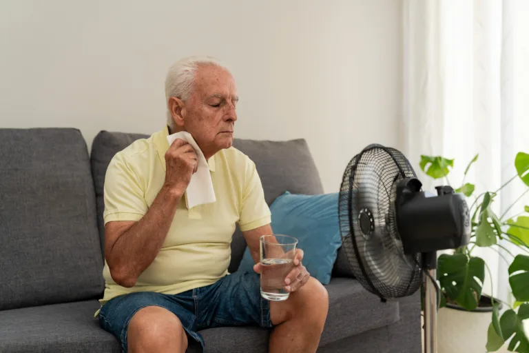 Elderly man refreshing himself with a glass of water and an electric fan, trying to cope with high temperature at home during summer heatwave