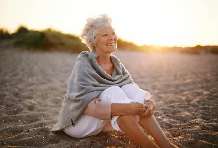 Happy retired woman wearing shawl sitting relaxed on sand at the beach. Senior caucasian woman sitting on the beach outdoors