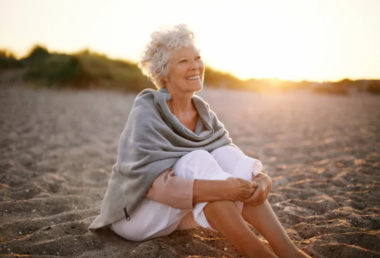 Happy retired woman wearing shawl sitting relaxed on sand at the beach. Senior caucasian woman sitting on the beach outdoors