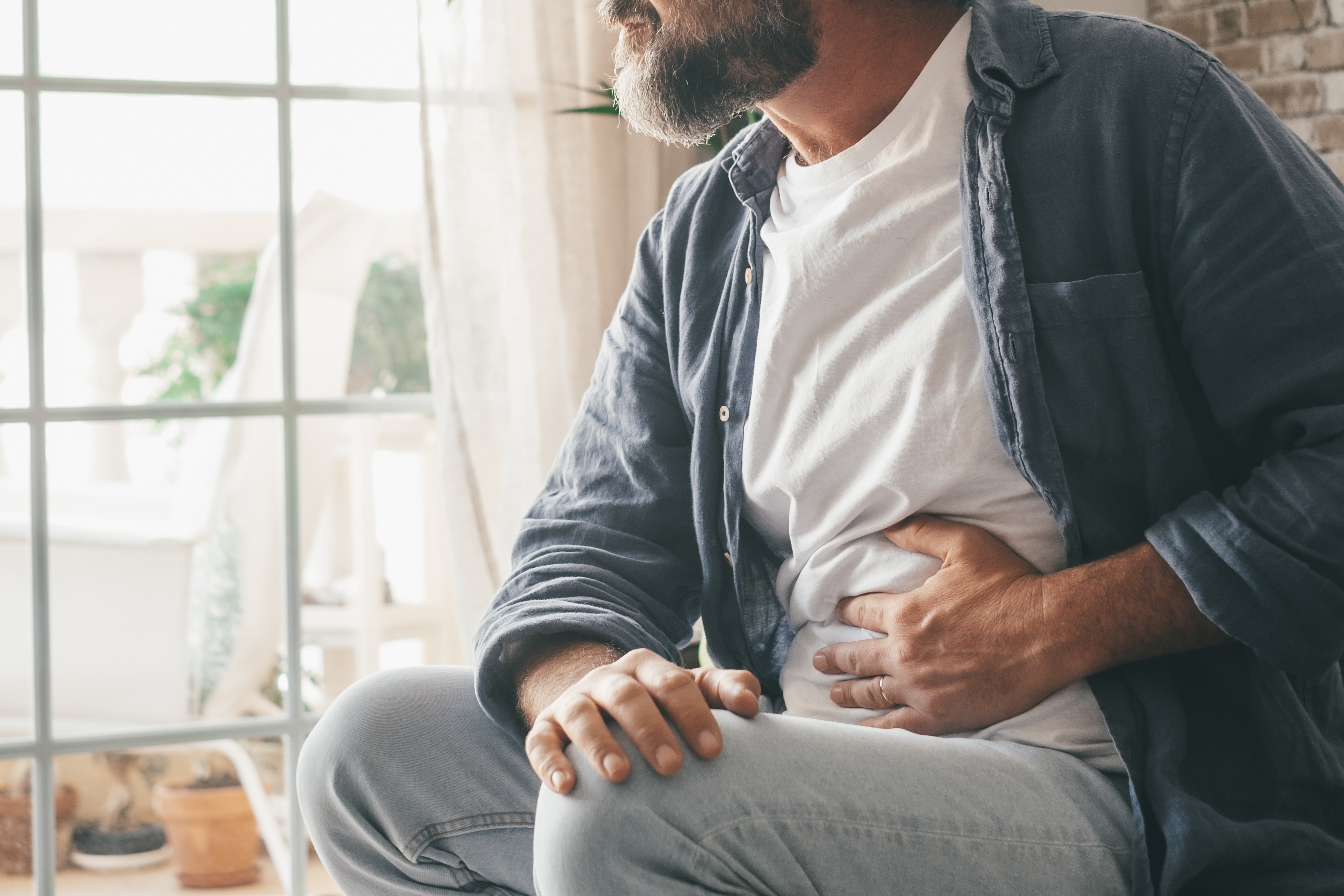 Zakaj boli trebuh? To so najpogostešji razlogi Man suffering with severe stomach pain sitting at home. Hand of mature guy holding abdomen suffering from ache, diarrhea or indigestive problem. Caucasian guy pressing on belly on painful sensation