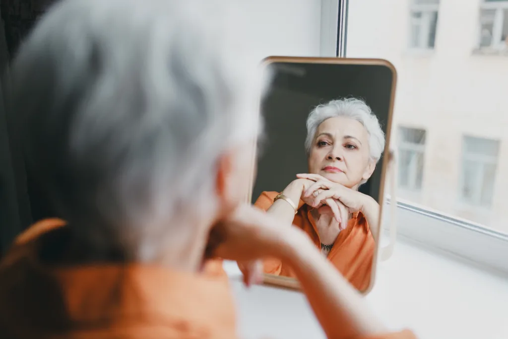 Senior elderly female with gray hair and wrinkles looking in mirror, upset with aging process of her body, reflecting upon her youth and young facial skin, missing her past beautiful appearance
