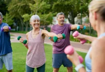 Za boljše ravnotežje, več moči in manj padcev: jeseni prihaja 194 brezplačnih vadb za odrasle Elderly group of people working out with dumbbells in a park. Mature fitness group of women and men lifting small weights outdoors. Happy active adults in sportswear exercising together with dumbbells.
