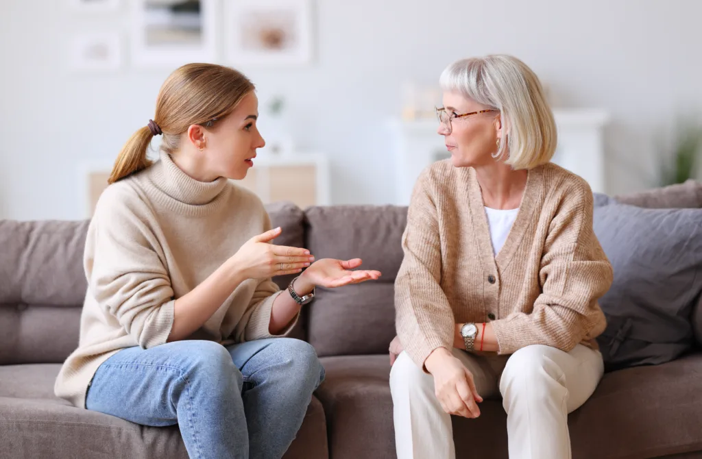 Adult daughter gesticulating and speaking with elderly mother while resting on couch at home together