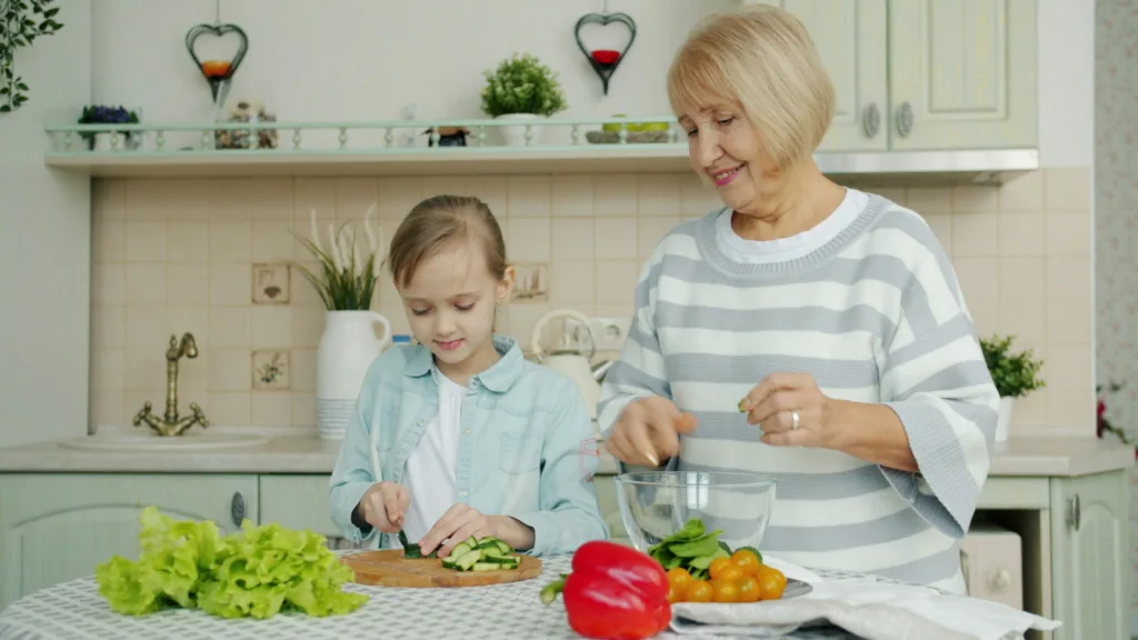 Granddaughter and grandmother are making salad together and talking in kitchen at home, little girl is cutting vegetables while old woman is watching her.