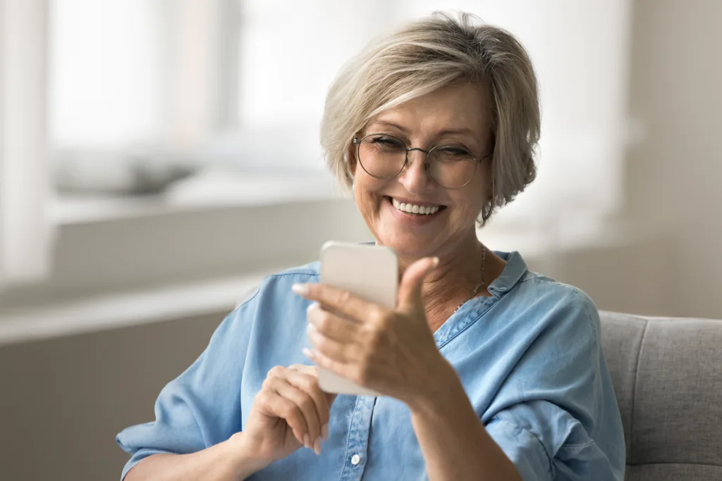 Older woman with short hair wearing glasses holding smartphone looking at screen with joyful expression, enjoy on-line chat with friend or family, making call to grown up children using modern device