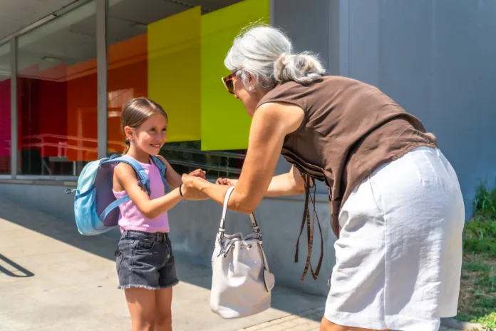 Grandmother bending down, happily greeting her smiling granddaughter with a backpack outside a colorful modern school building, symbolizing family support and intergenerational bonding