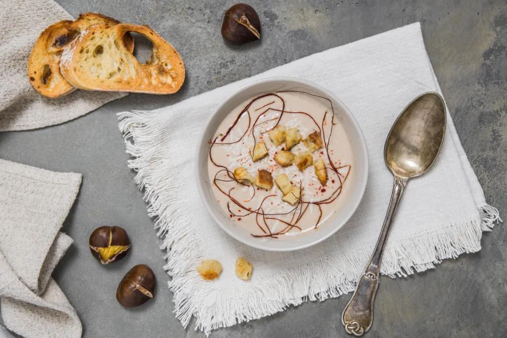 Top view on chestnut soup with parmesan, croutons and chilli flakes and threads on grey concrete background, decorated with roasted bread and chestnuts, top view