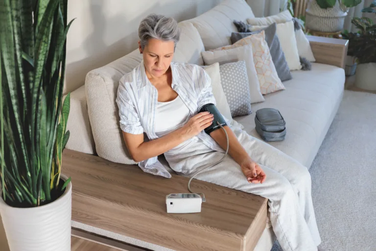 Older woman sitting on a sofa, using a digital blood pressure monitor. Focused on her health, she checks vital signs in a cozy home environment. Aging with responsibility and care.