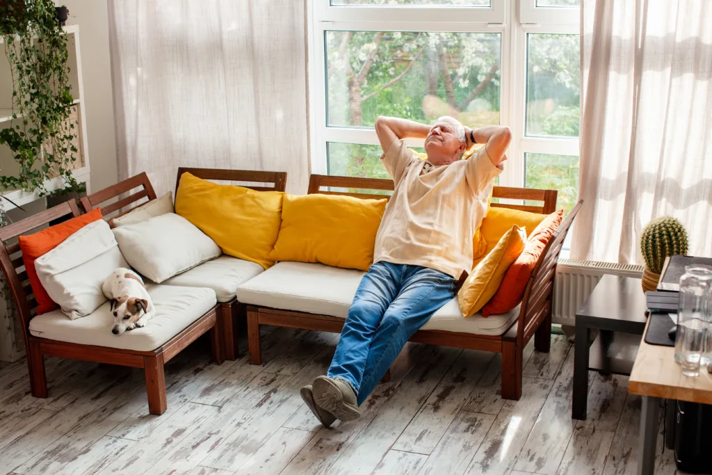 Senior man relaxing on a wooden sofa with his dog in a bright living room. Retired grandfather resting at home after work. Concept of peaceful retirement, comfort, and cozy leisure.